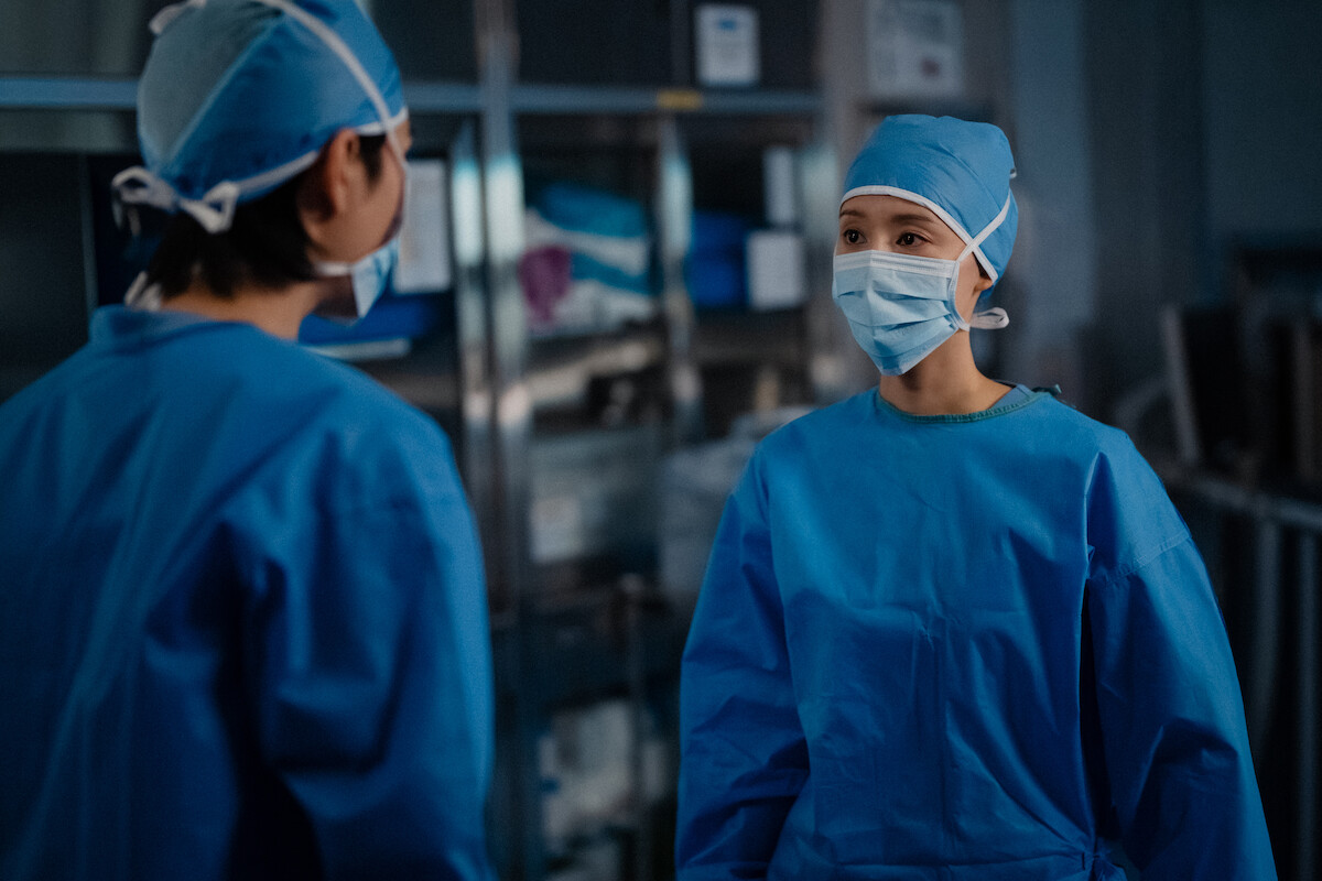 Duas pessoas asiáticas se olham vestidas com trajes médicos típicos de uma sala de cirurgia