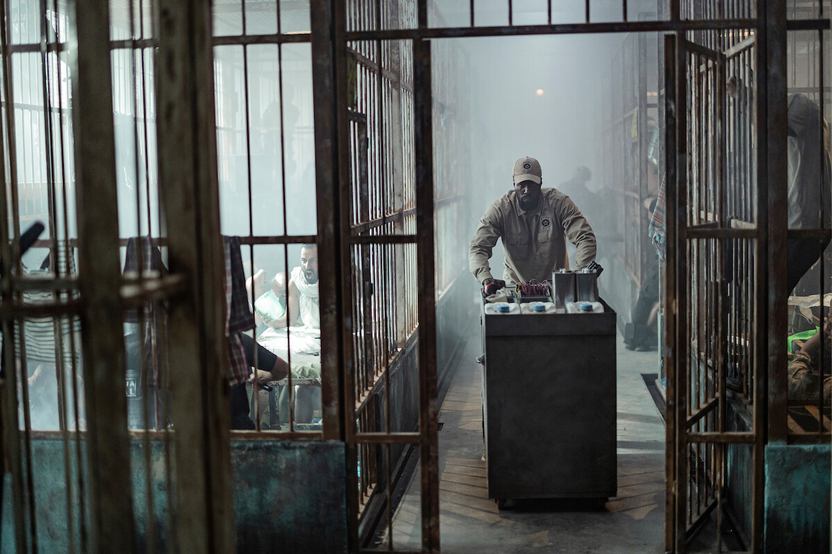 A prison scene with a guard pushing a food cart down a cell-lined corridor, while inmates sit inside their cells in a dim, smoky environment.