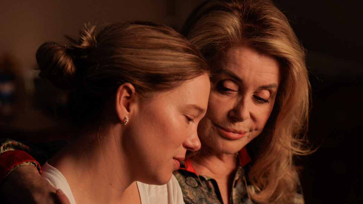 Two women sit close together in a warmly lit room, one older with an arm around the younger, both sharing a quiet, intimate, and comforting moment, suggesting a deep emotional bond or support in a private setting.