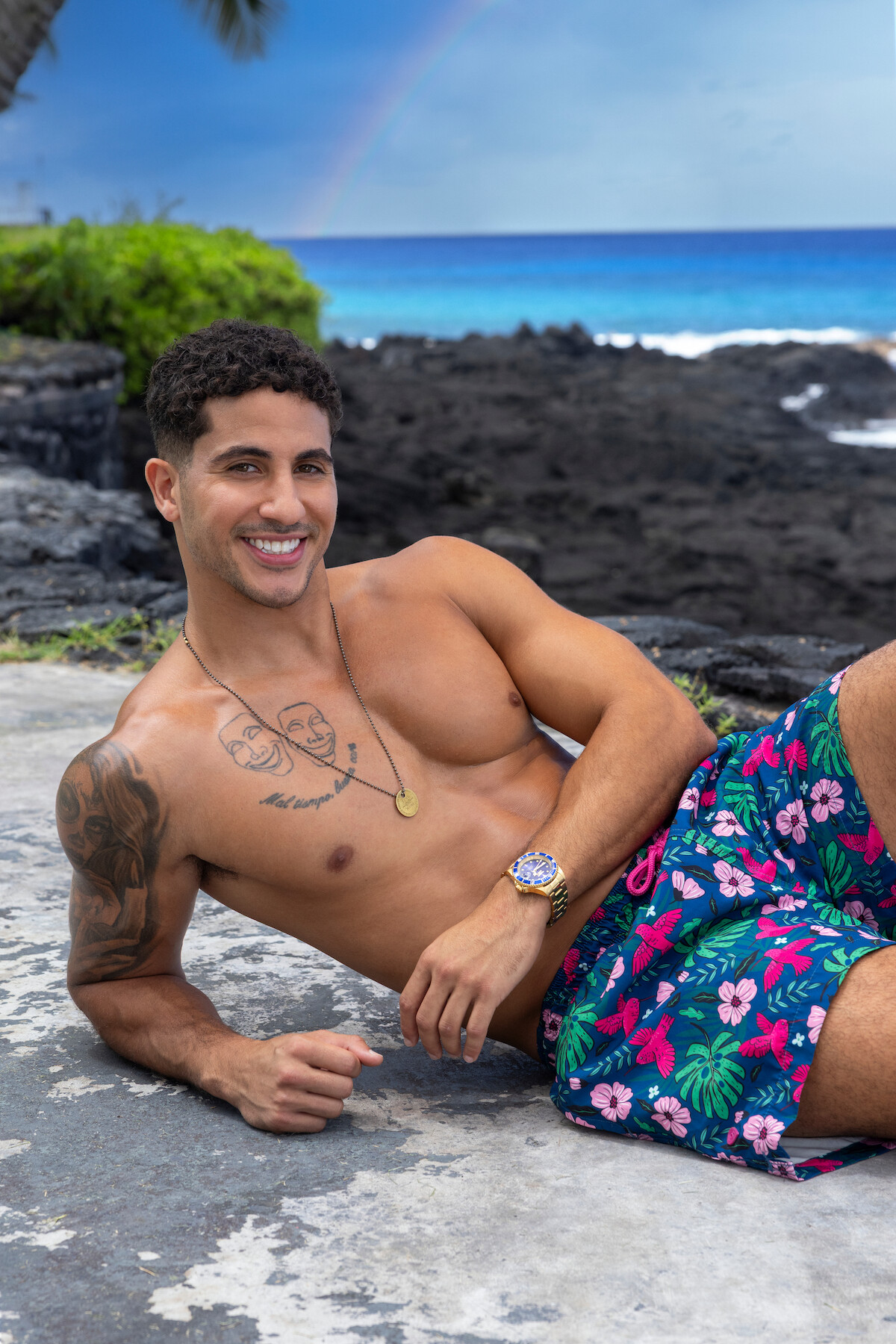 Smiling shirtless man with tattoos and floral swim trunks relaxes on rocky shore by ocean, with lush greenery, a palm tree, and a faint rainbow in the background under a blue sky.