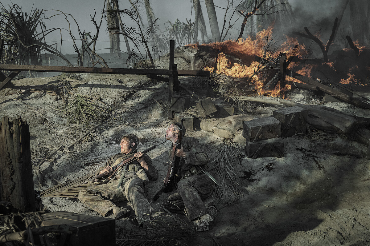 Two soldiers in dirty uniforms lie on a smoky, war-torn battlefield holding rifles, surrounded by debris and crates, as fire rages behind them and palm trees stand amid destruction.