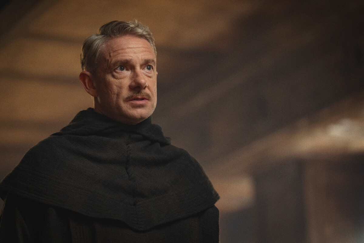 A man wearing a black robe stands indoors in a dimly lit setting with wooden beams, looking slightly upward with a serious expression, suggesting a historical or monastic environment.