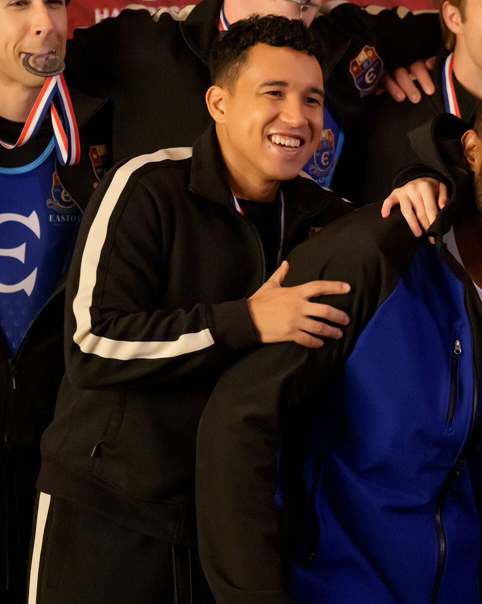 Smiling young man in a black tracksuit with a white stripe stands with teammates, some wearing medals, in a celebratory indoor team photo setting.