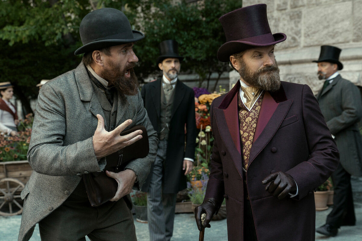Two men in Victorian-era attire converse outdoors; one gestures animatedly while the other listens holding a cane. Background shows other well-dressed men, potted flowers, and stone building, creating a historical setting.