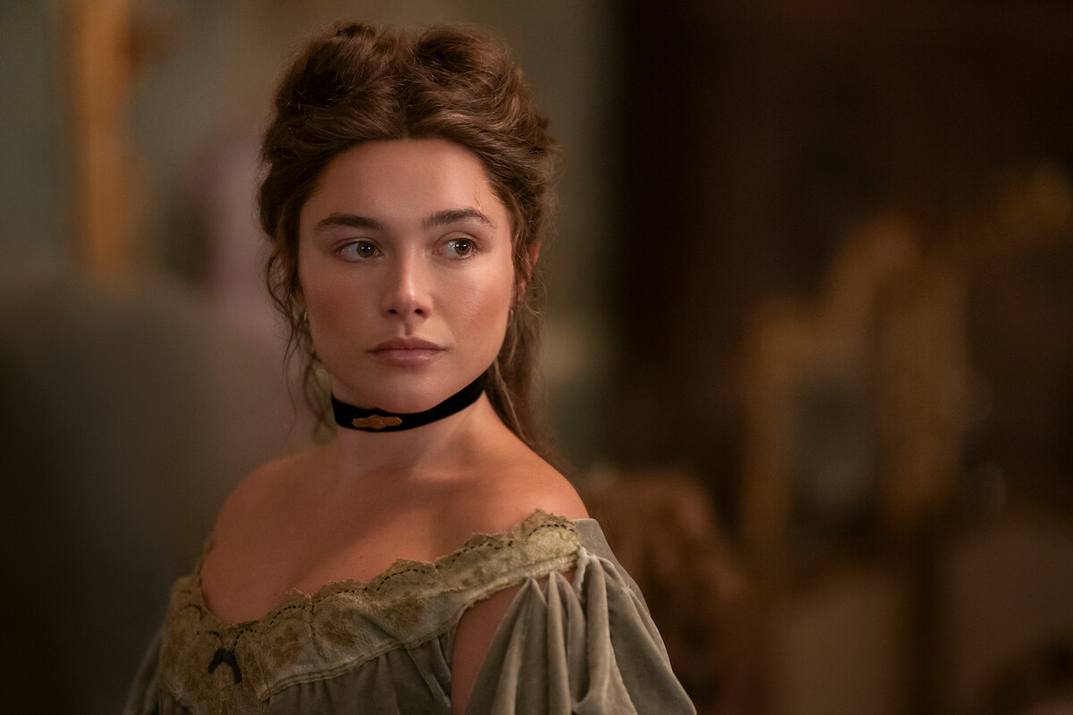 Woman in vintage clothing with off-shoulder dress and black choker stands indoors, soft lighting, blurred ornate background, calm and thoughtful expression.