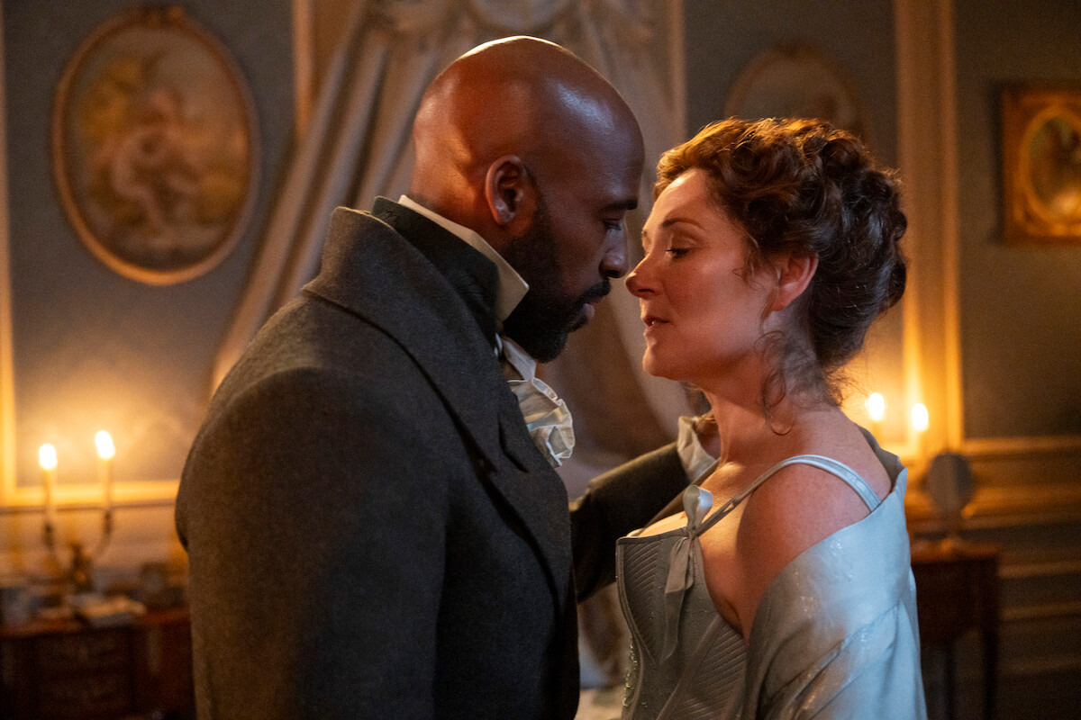 A man and woman in period clothing stand closely, gazing into each other’s eyes in a candlelit, ornately decorated room with framed paintings and draped curtains, creating a romantic atmosphere.