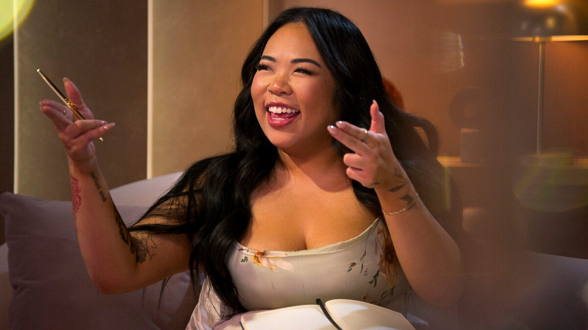 A woman with long dark hair smiling and gesturing enthusiastically while sitting indoors in a cozy, warmly lit room, holding a pen and notebook, creating a lively and positive atmosphere.