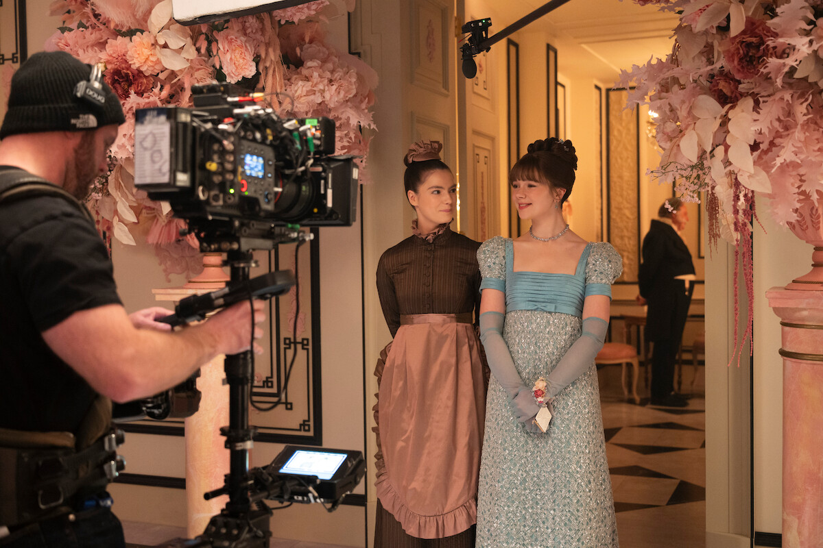 Duas mulheres com figurinos de época estão de pé em um corredor elegante, decorado com flores, em um set de filmagem, enquanto são filmadas em um momento de bastidor de uma produção histórica.