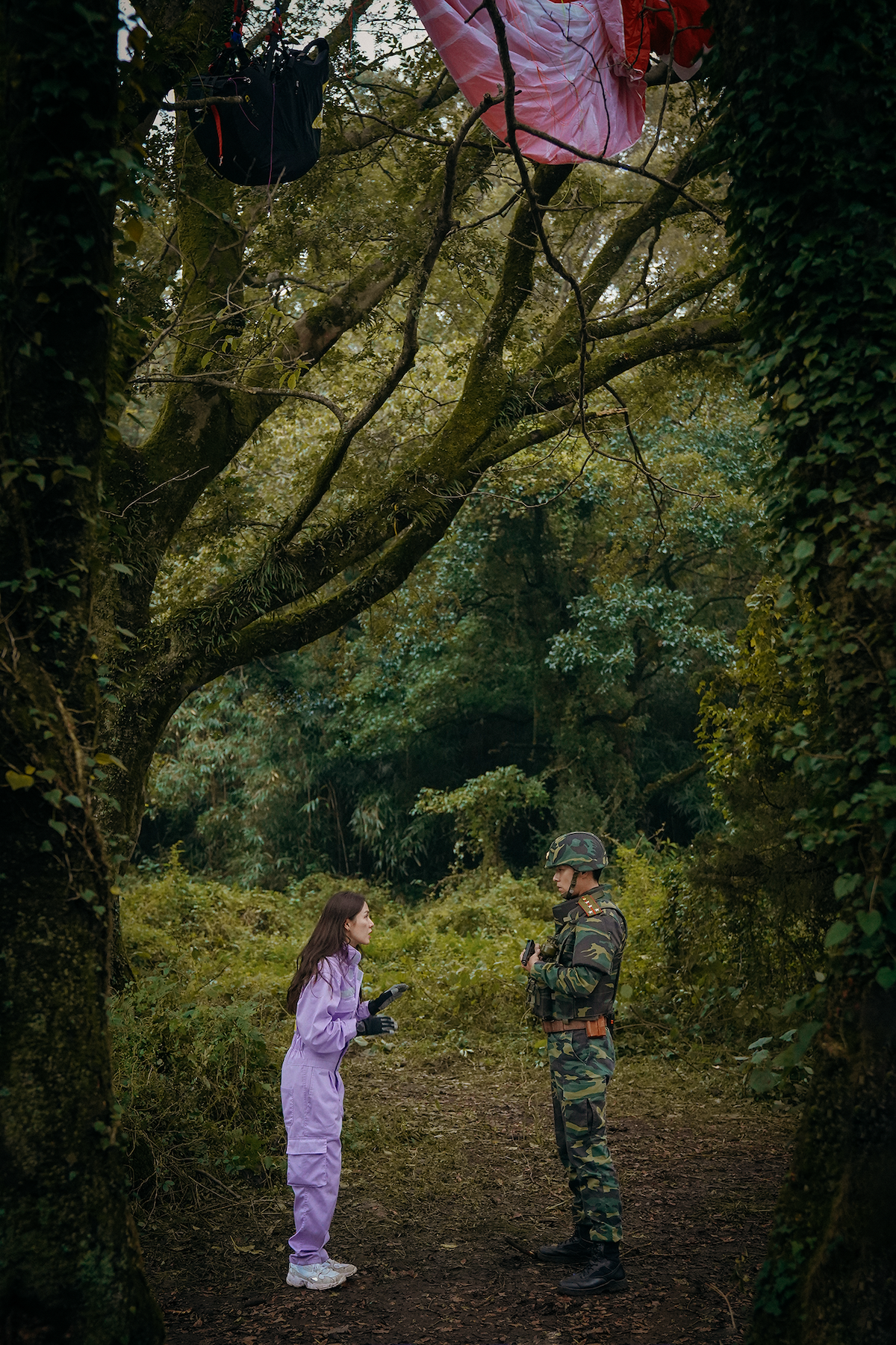 Um homem em trajes militares e uma mulher de macacão roxo conversam no meio de uma floresta, com um parapente preso a uma das árvores próximas