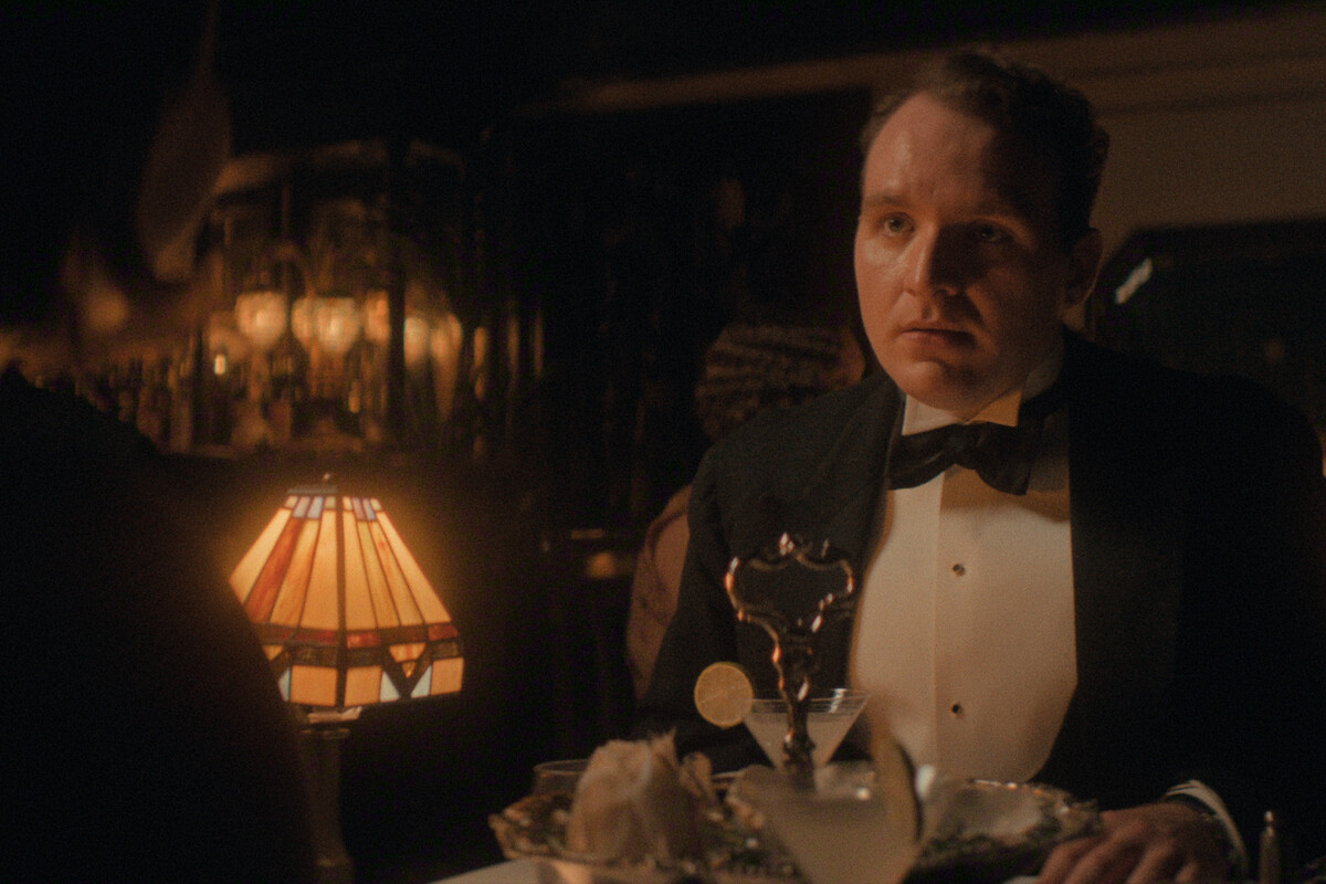 A man in formal wear sits at a candlelit restaurant table with cocktails and dinnerware, facing another person whose back is visible. The setting is elegant, warm, and dimly lit, with a vintage lamp providing soft light.