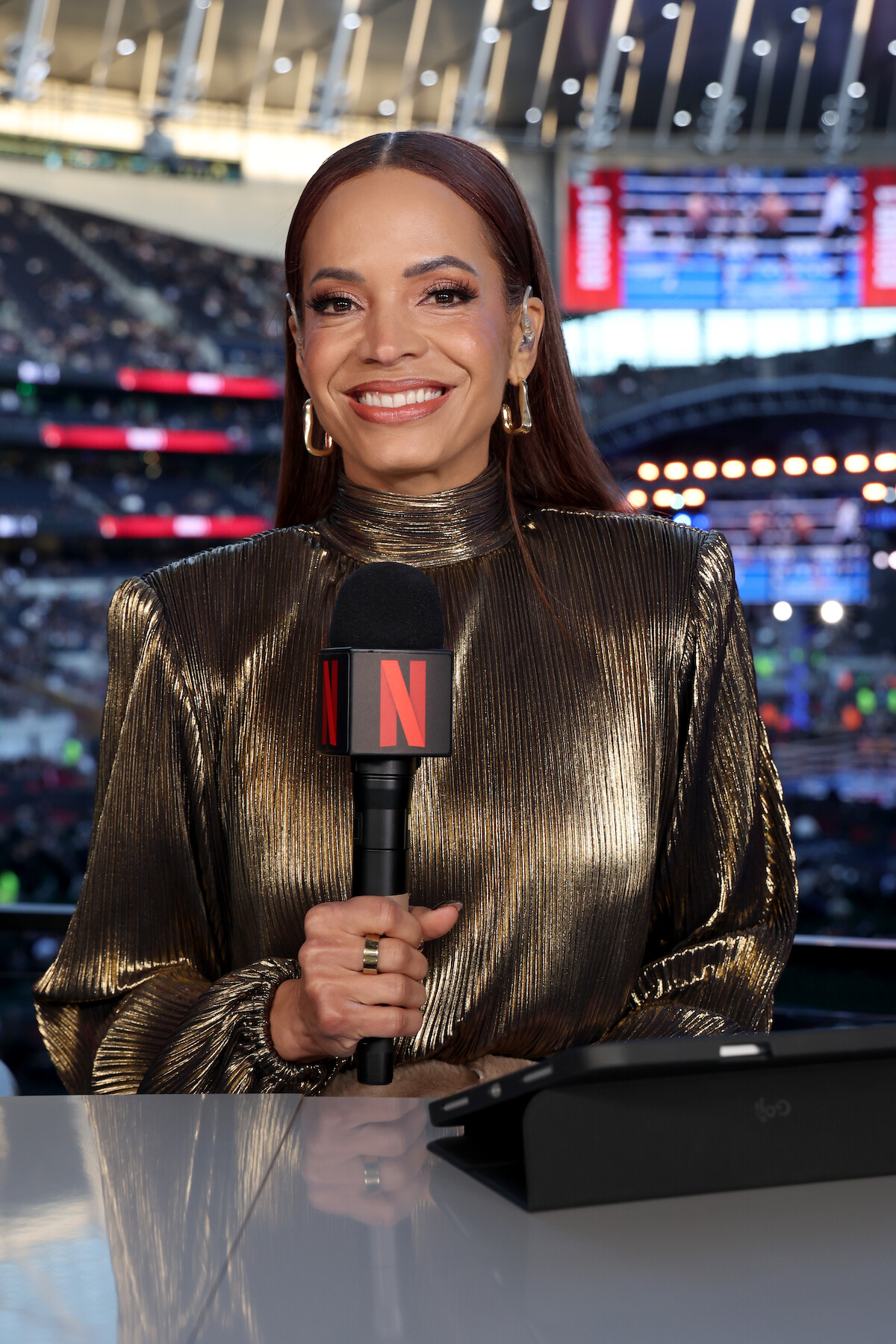 Woman in metallic dress holding Netflix microphone, smiling at a desk in a stadium filled with people and bright lights, with a large screen and arched roof structure in the background.