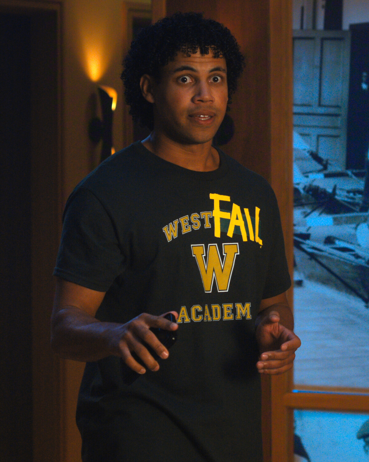 Man with curly hair wearing a "West Fail Academy" t-shirt stands indoors, looking surprised or startled, holding a remote control, with dim lighting and a cluttered room visible in the background.