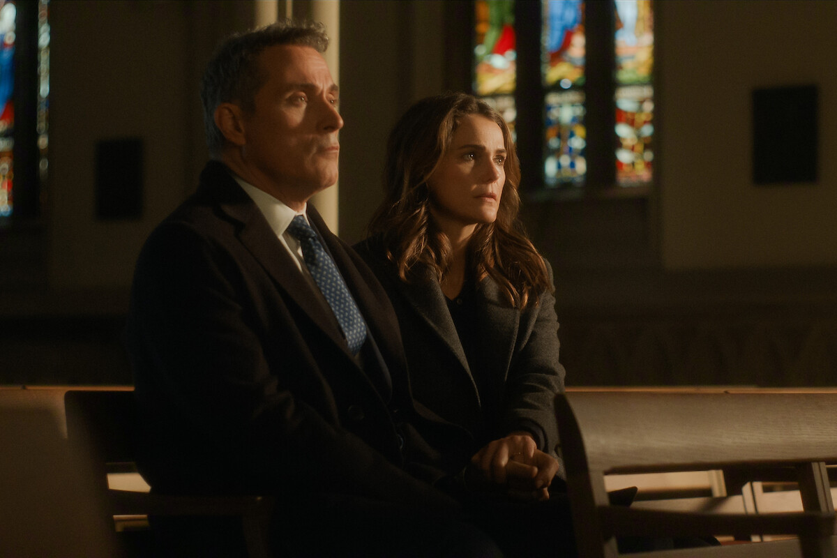 Two people sit closely on a church pew, holding hands, with soft light from stained glass windows in the background, suggesting a quiet, contemplative or emotional moment in a solemn church setting.