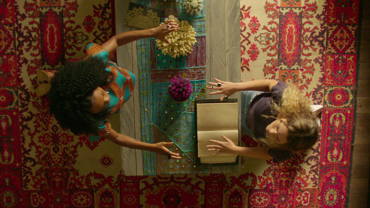 Two women sitting across from each other at a decorated table with flowers and an open book, viewed from above, on a patterned red carpet in a cozy indoor setting.