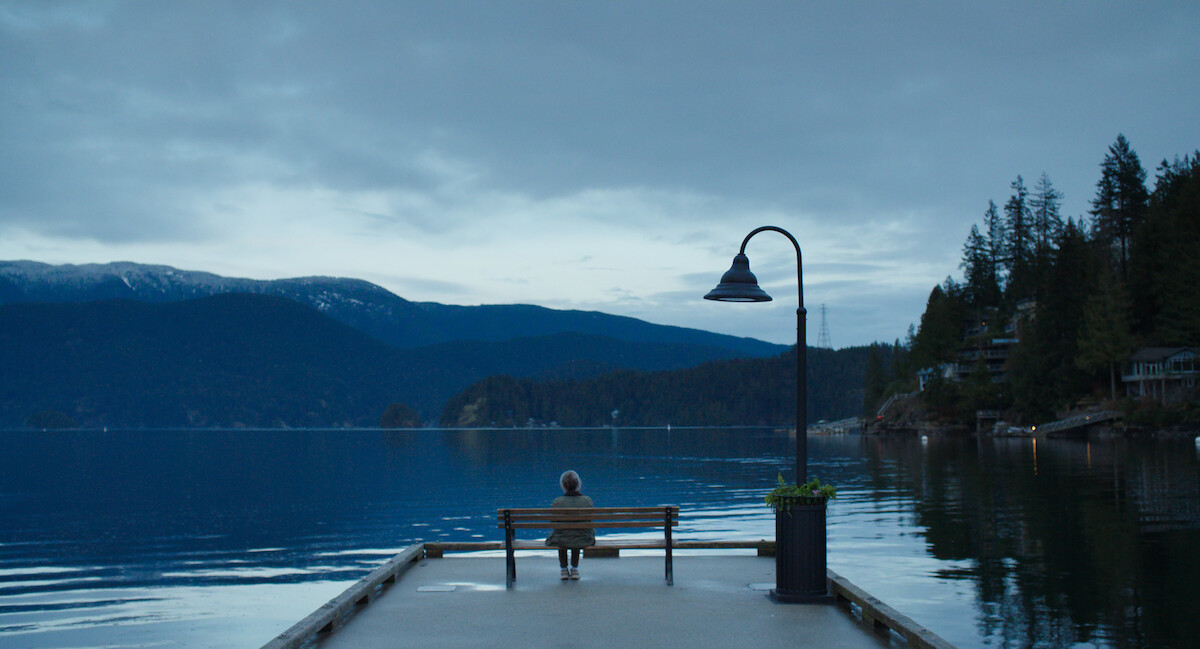 Sally Field as Tova sitting alone on a bench at the end of a dock overlooking a calm lake, surrounded by forests and mountains under a cloudy sky, with a lamppost beside them and houses visible along the shoreline.