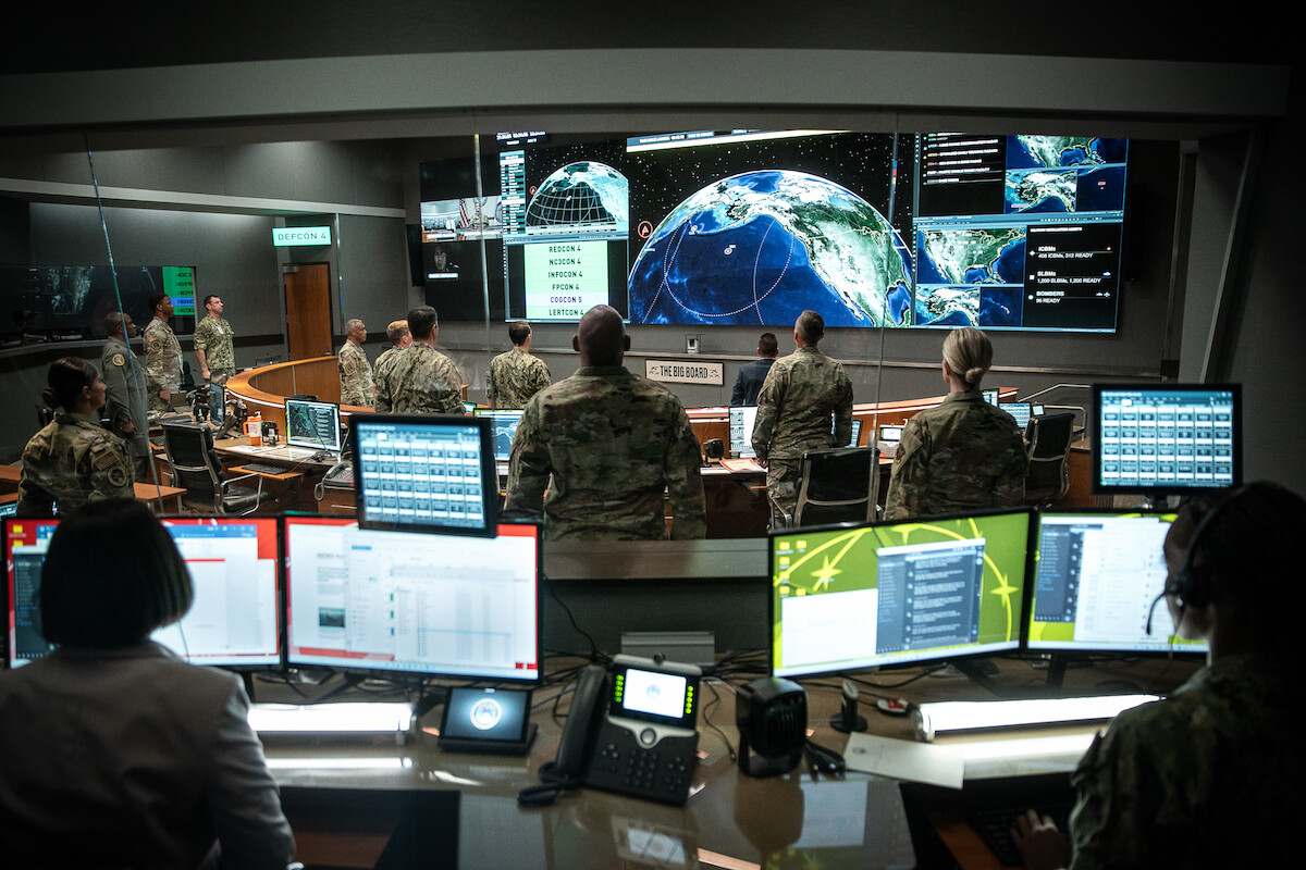 Military personnel in a high-tech control room monitor multiple screens with satellite and earth data. The atmosphere is serious and focused, with rows of computers and large digital displays showing global strategic information.
