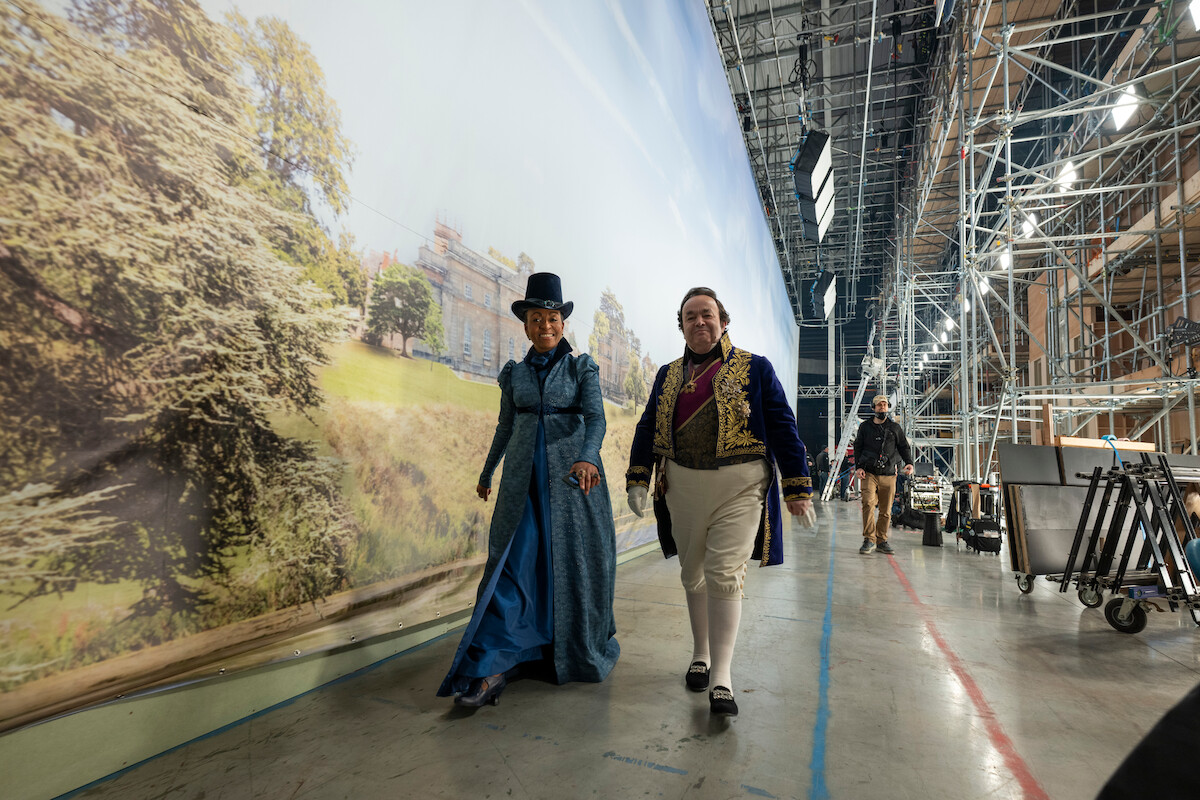 Duas pessoas com figurinos de época caminham por um set de filmagem com andaimes e equipamentos, ao lado de um grande painel pintado que retrata uma paisagem ao ar livre com árvores e uma mansão.