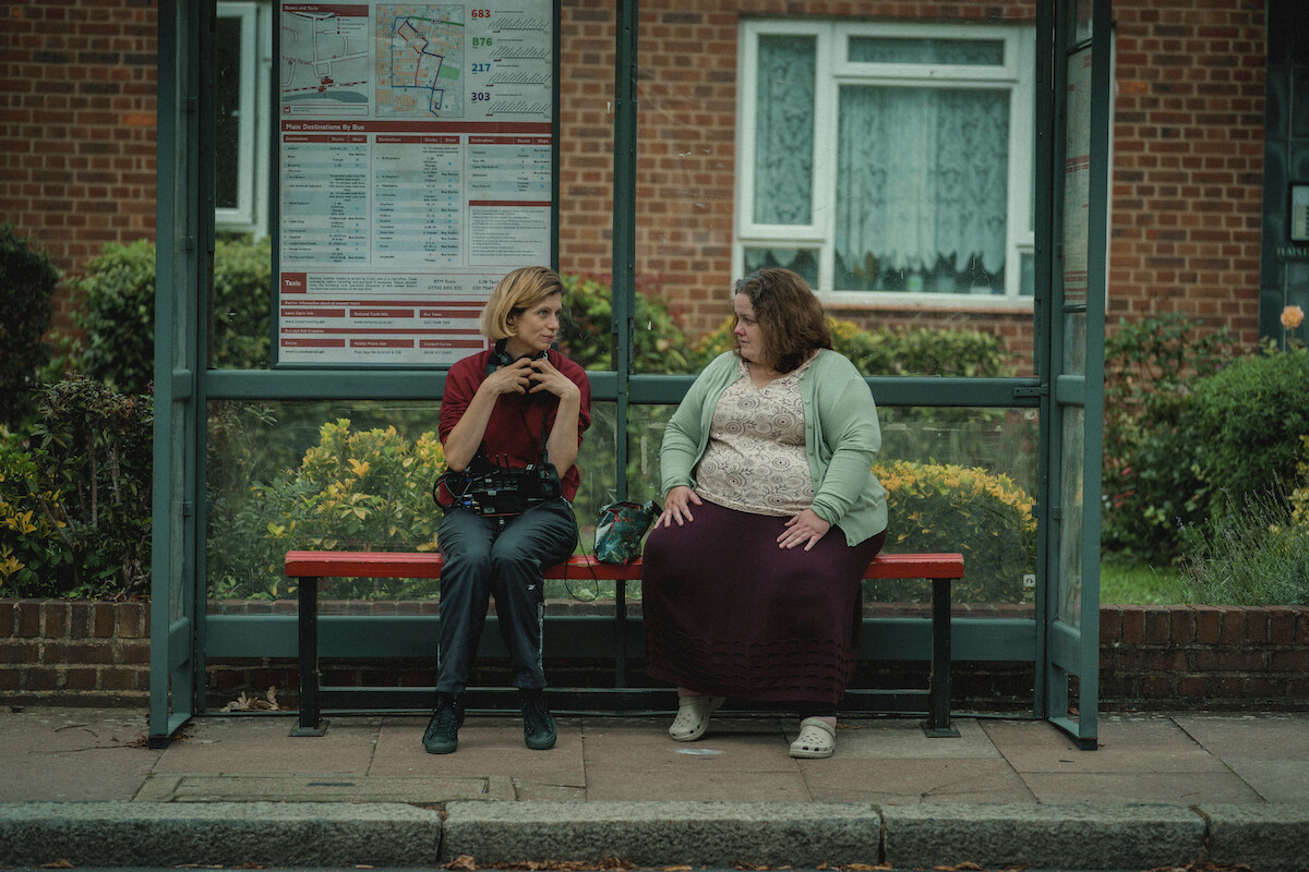 Weronika Tofilska and Jessica Gunning sit on a bus bench.