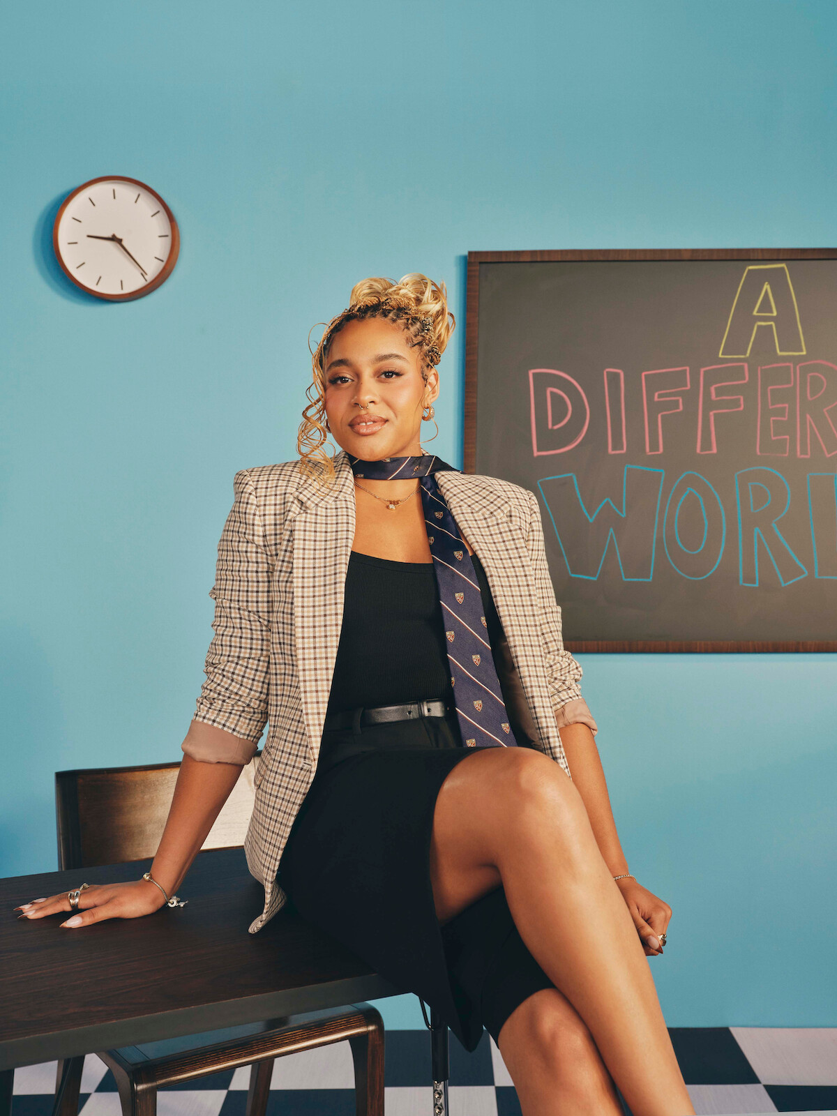 Woman sitting on a desk in a classroom, wearing a blazer, dress, and tie, with a chalkboard that says "A Different World" and a clock on a blue wall in the background.