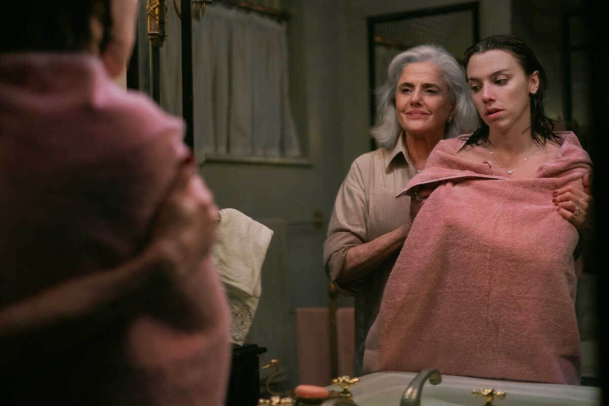 Duas mulheres se abraçam em um banheiro. A primeira está molhada e enrolada em uma toalha rosa. A segunda a abraça carinhosamente por trás e esboça um sorriso. Ela tem os cabelos grisalhos e veste camisa marrom