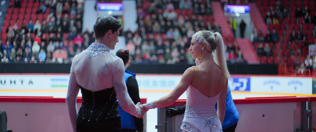 Paul Poirier and Piper Gilles in costumes hold hands at the edge of an indoor ice rink, preparing to perform while an audience watches from the stands in a large arena.