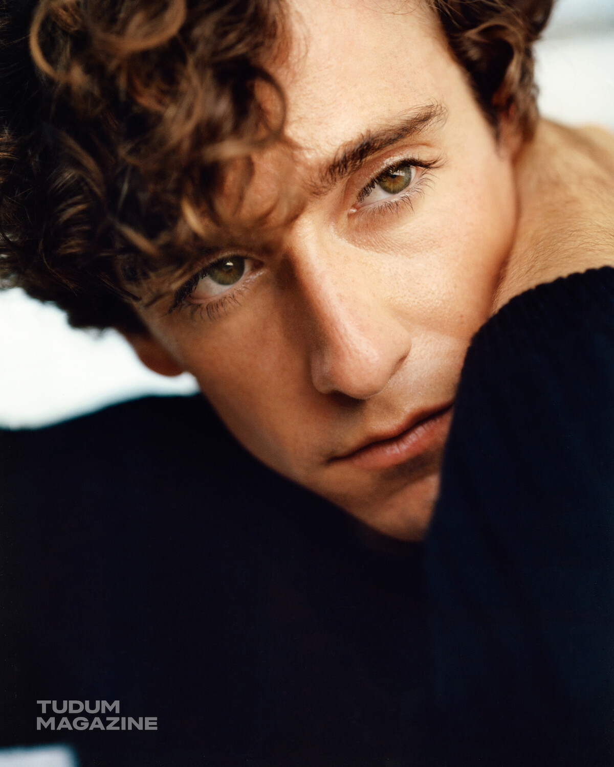 Close-up of a young person with curly hair and green eyes, gazing thoughtfully into the distance while resting their chin on their hand, wearing a dark sweater in a softly lit indoor setting, with a Tudum Magazine watermark.