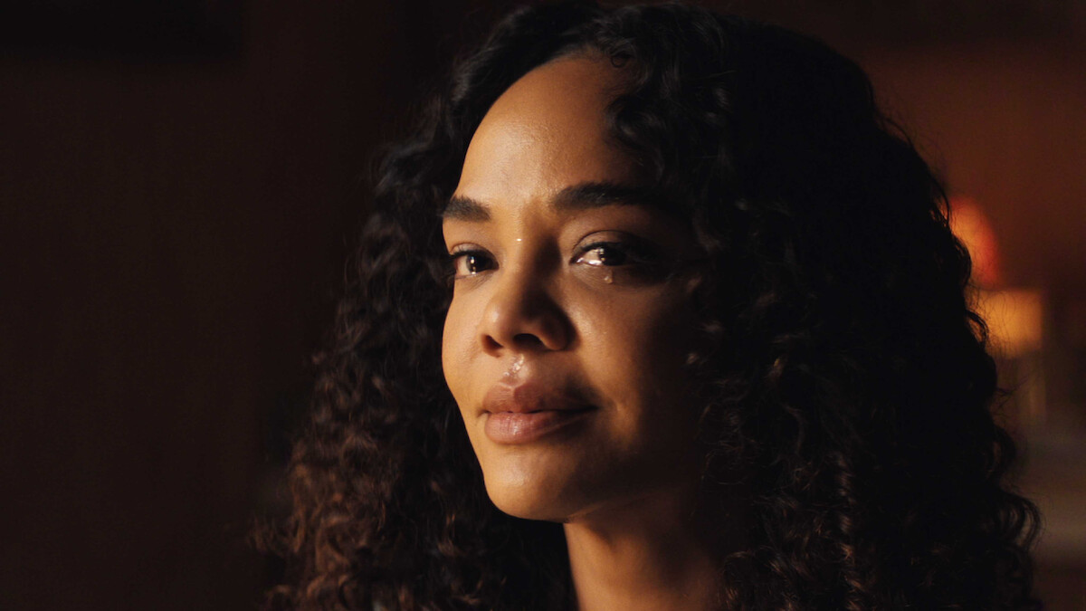 Woman with curly hair in a warmly lit indoor setting, looking slightly off-camera with a calm and introspective expression, soft background lighting suggesting a cozy and intimate environment.