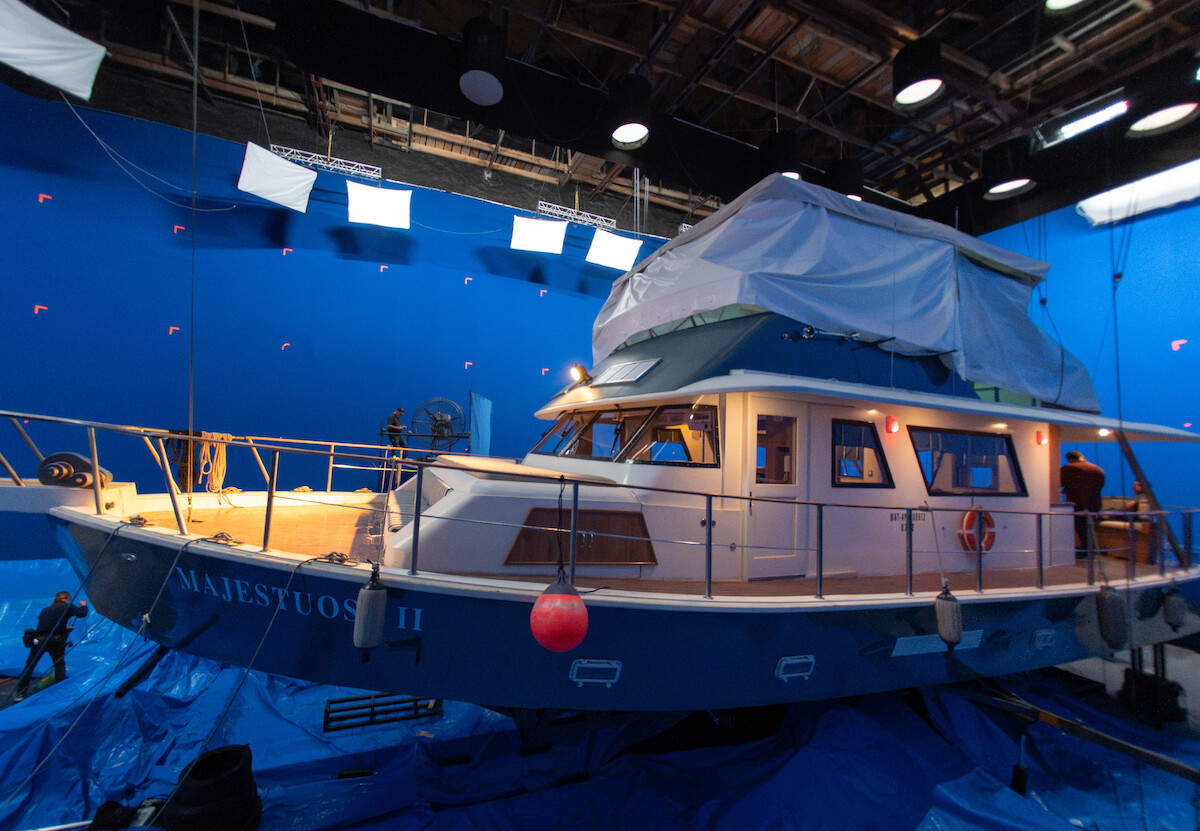 Um grande barco pintado nas cores azul e branco está no meio de um set de filmagem, cercado por pedaços de lona e holofotes de iluminação