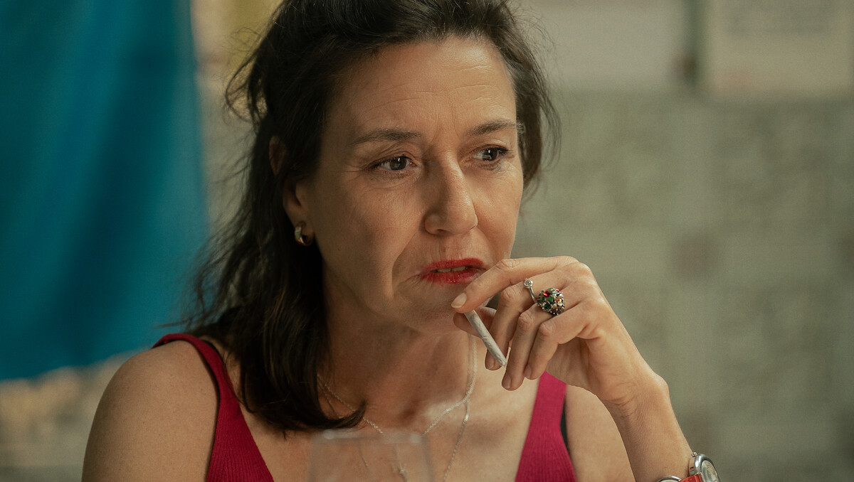 Middle-aged woman with dark hair in a red top holding a cigarette, thoughtfully gazing to the side indoors; soft lighting, blurred background with neutral tones and a hint of blue.