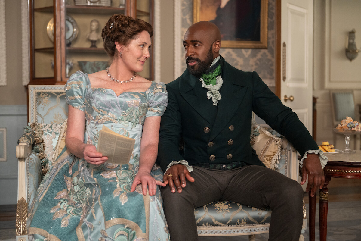 A woman in a floral Regency-era dress and a man in period costume sit on an ornate sofa in an elegant, historical room, engaged in conversation.
