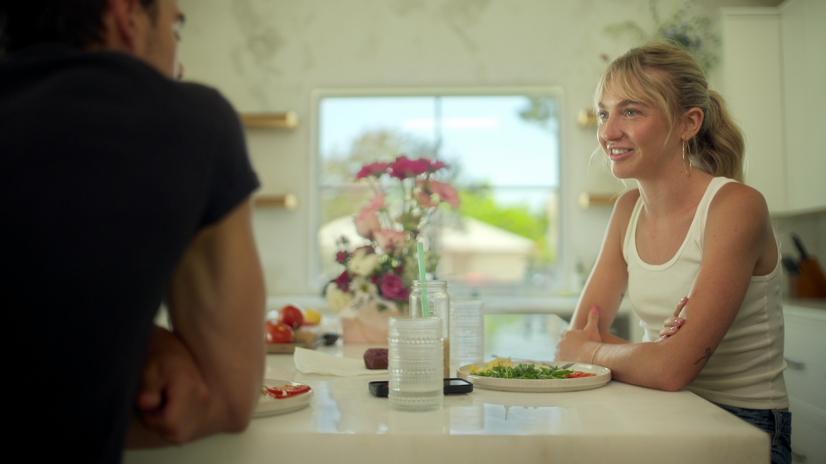 Two people sit at a kitchen counter with plates of food, glasses, and flowers on display. They are talking in a bright, modern kitchen with sunlight coming through a window.