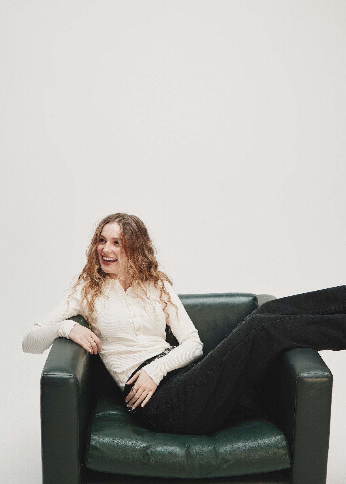 A smiling woman with long hair sits casually on a dark armchair, wearing a white long-sleeve top and black pants, with one leg draped over the armrest, in a minimalist studio with a light background.