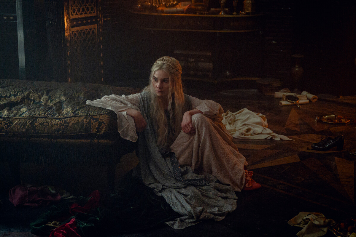 A woman in a long, pale dress sits pensively on the floor next to a decorative sofa in a dim, cluttered, medieval-style room, evoking a somber, reflective mood. Scattered clothes and low lighting add to the atmosphere.