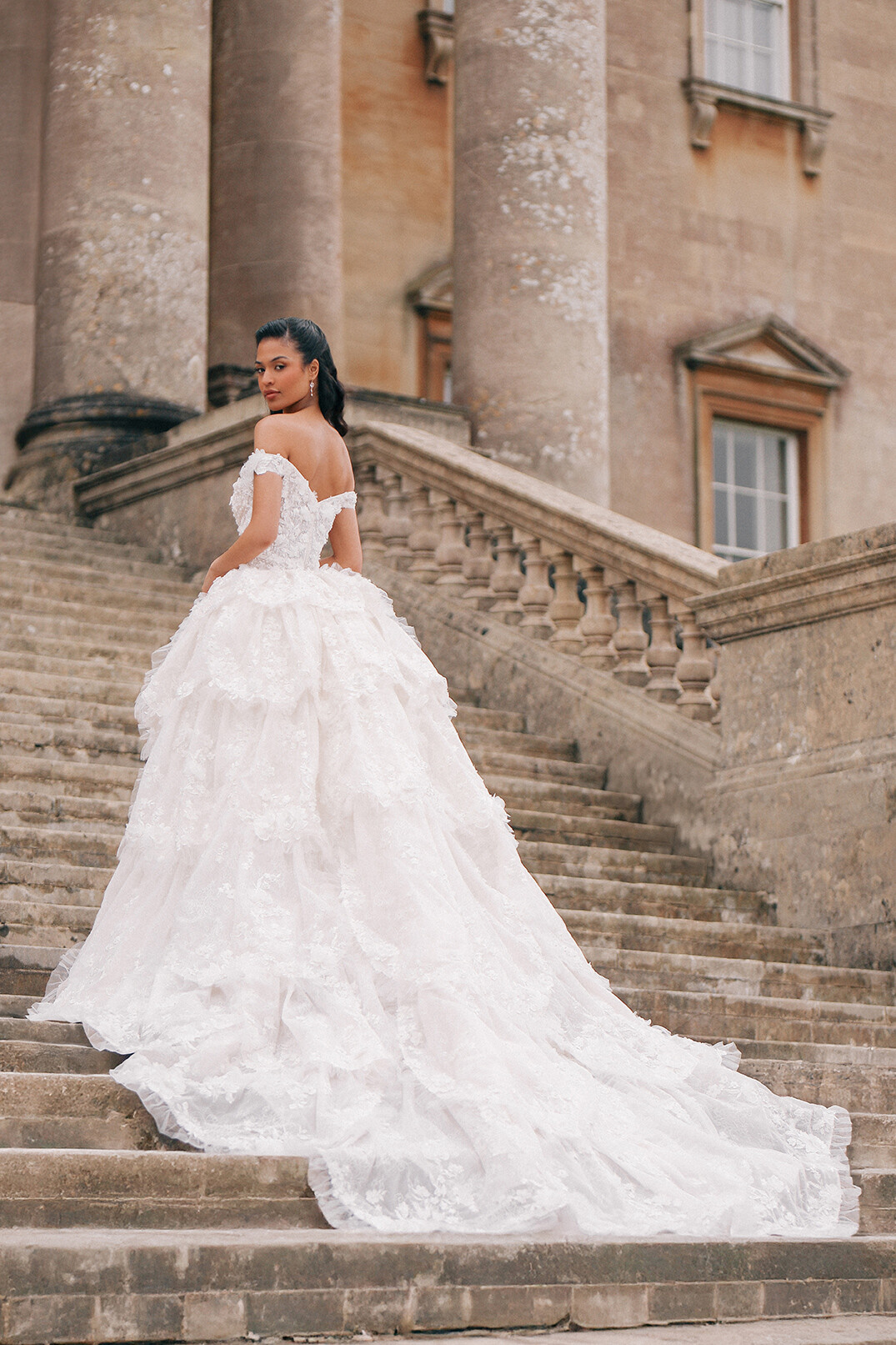 A bride in an elegant white gown stands on grand stone steps outside a historic building, looking back over her shoulder; the mood is regal and romantic.