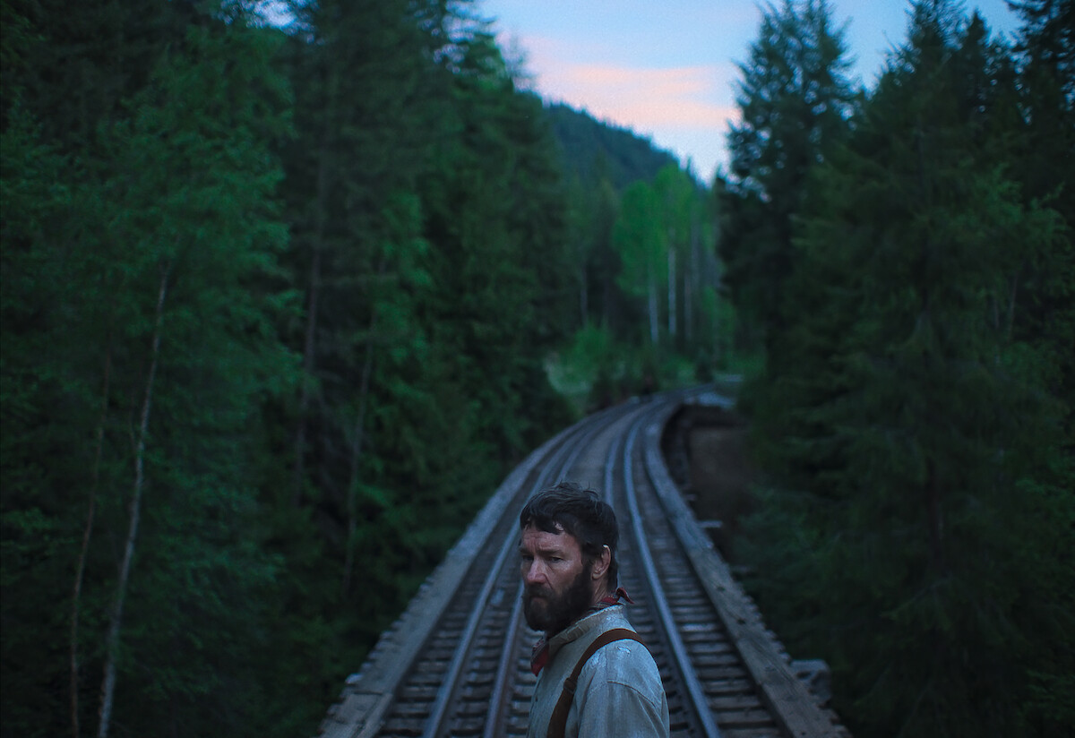 Joel Edgerton as Robert Grainier standing on railroad tracks that cut through the forest.