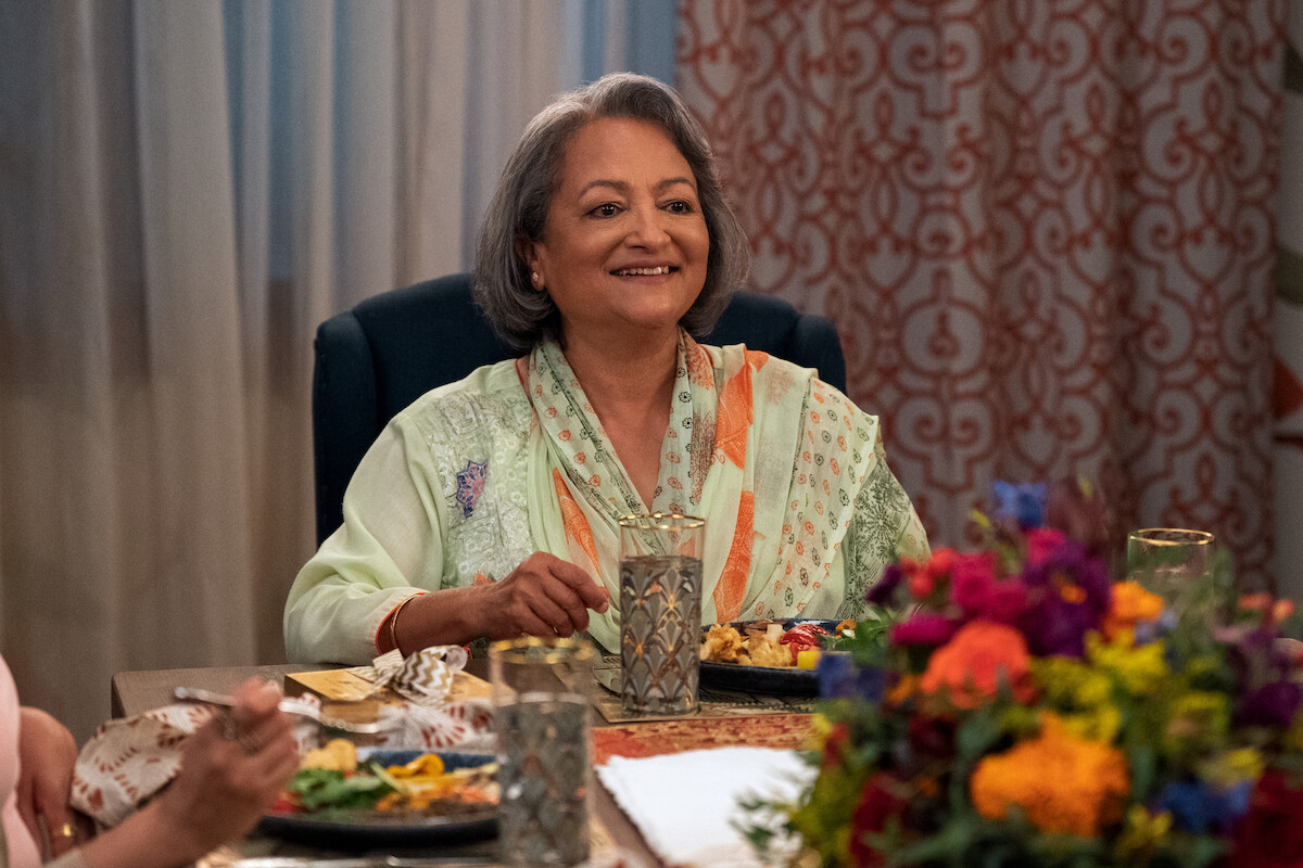 Ranjita Chakravarty as Nirmala sits at a table smiling in Season 4 of ‘Never Have I Ever.’ 