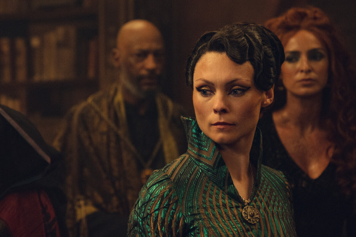 A woman in ornate, green, fantasy clothing stands in a dimly lit room with bookshelves. Other people in elaborate costumes are behind her. The atmosphere is tense and dramatic.