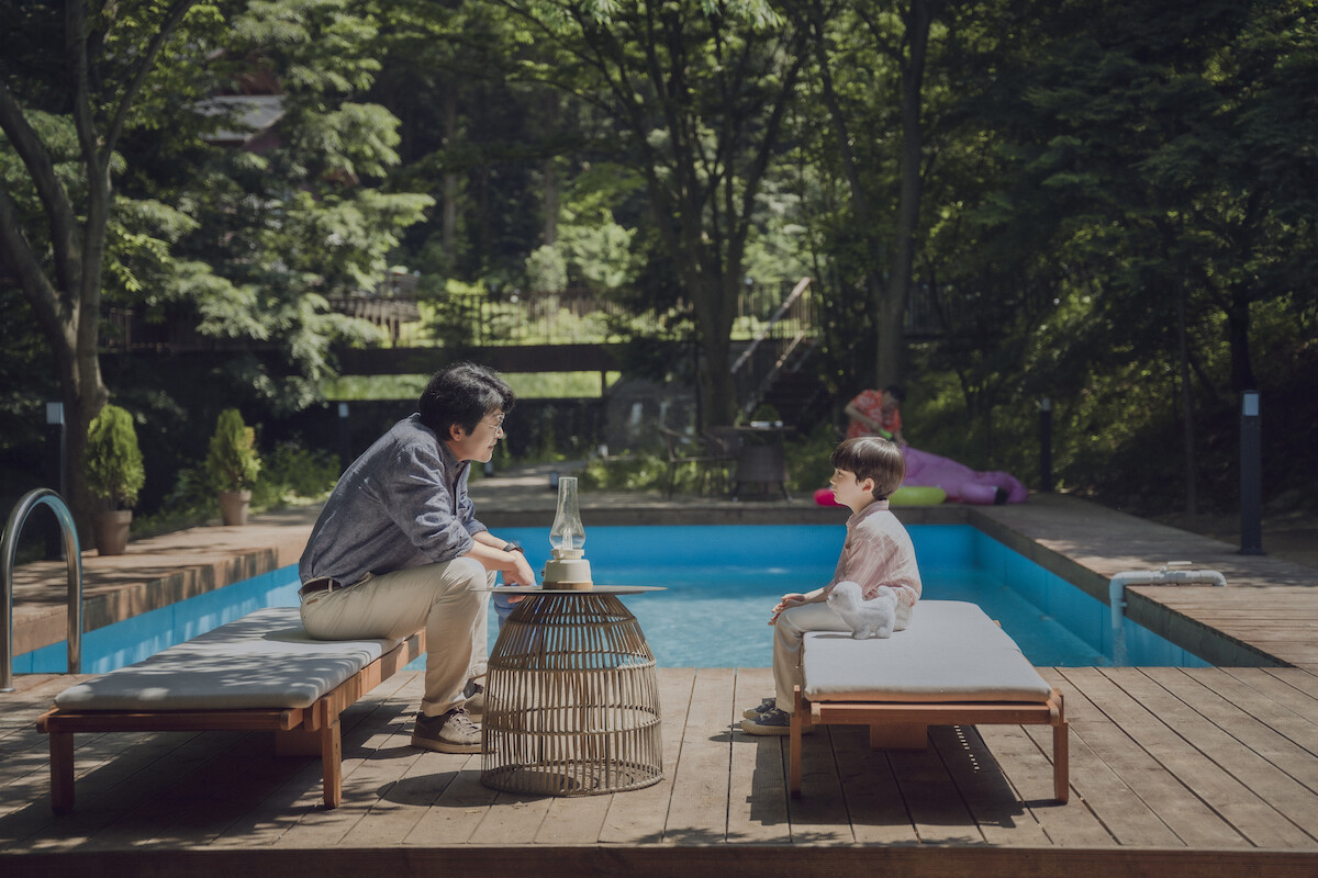 Kim Yun-seok as Jeon Young-ha and Jo Yeo-joon as Ha Si-hyeon sit across from each other on pool lounge chairs in front of an in-ground pool in an image from ‘The Frog.’