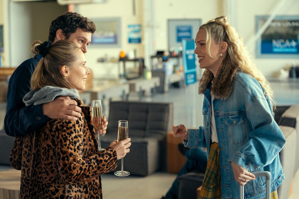 Three people talking and smiling in a bright, modern lounge. A couple holds champagne glasses while interacting with a woman in a denim jacket, who stands beside a suitcase. The setting appears to be an airport or travel lounge.