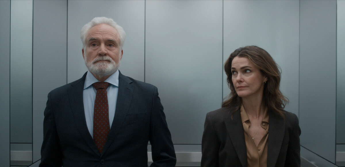 Two professionally dressed adults, a man and a woman, stand side by side in a modern, silver elevator. The woman glances at the man, both appear serious, elevator walls and lighting provide a sleek and formal office setting.