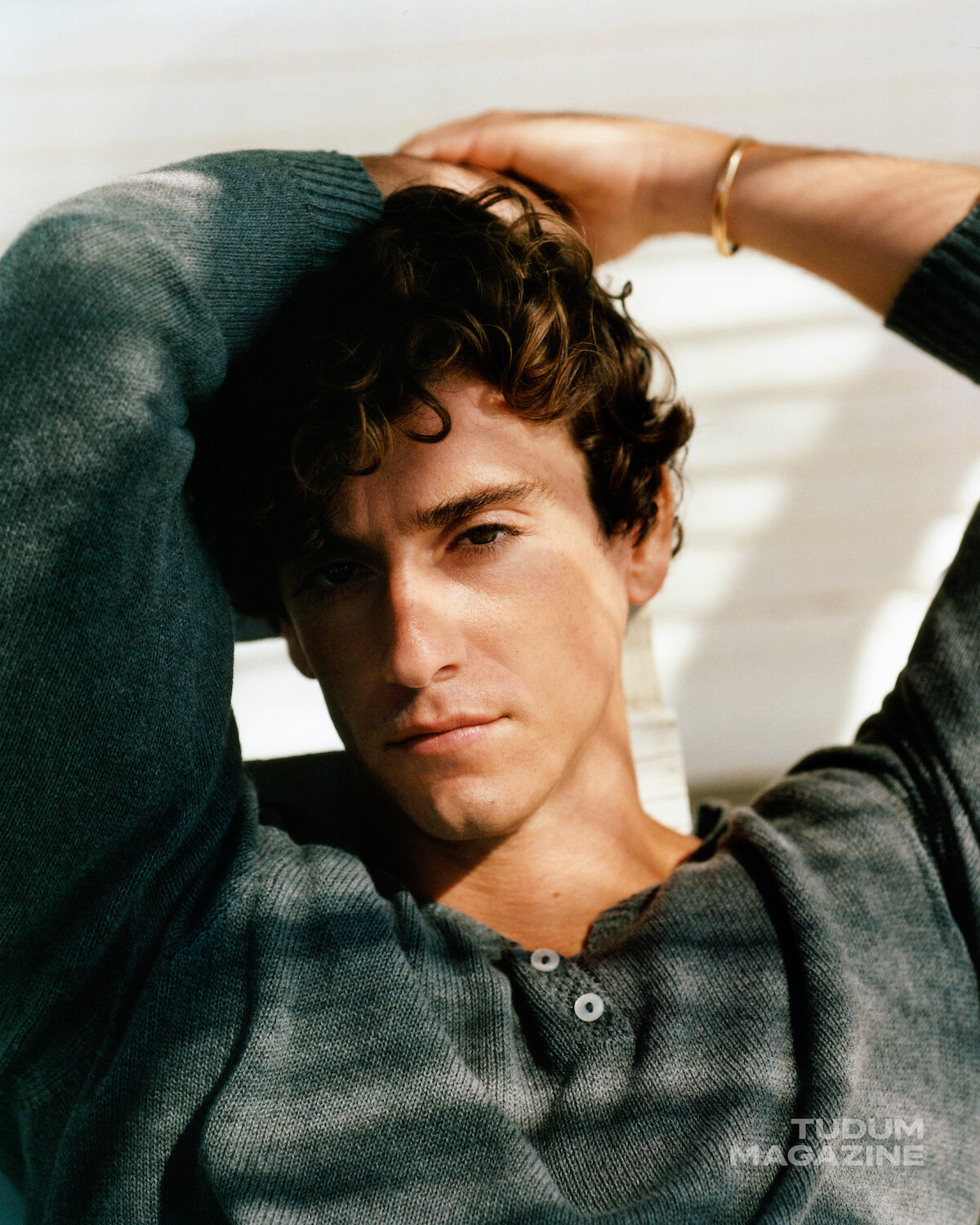 Young man with curly hair in casual sweater sits indoors, leaning back with arms raised behind his head. Sunlight filters through blinds, casting shadows across his face and body. Calm, contemplative atmosphere.