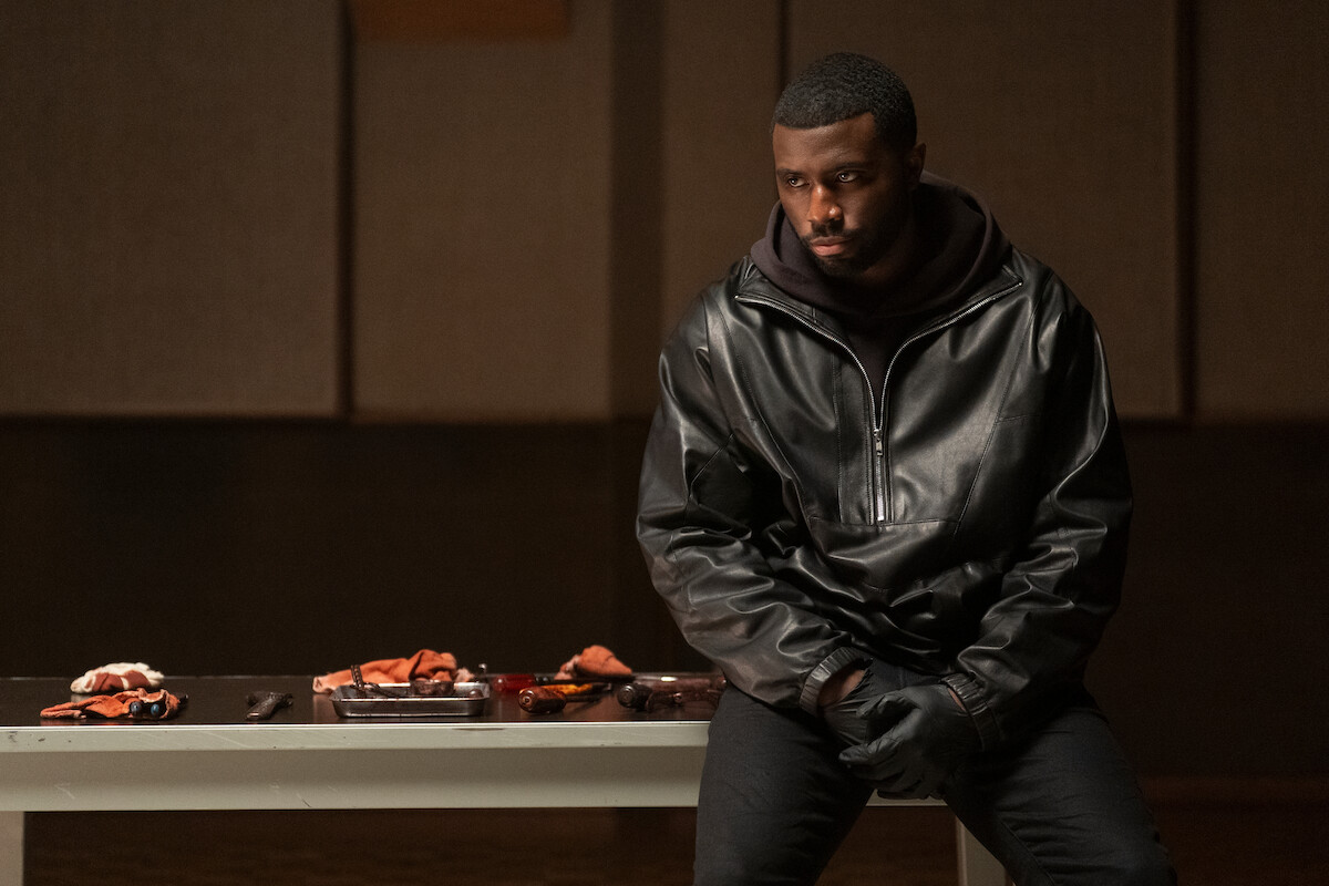A man in a black leather jacket sits on the edge of a white table indoors, with gloves on, looking serious. Assorted objects and food are spread on the table beside him. The room has soundproofed panels on the wall.