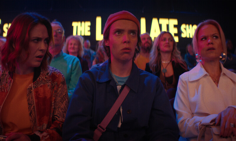 Three people sit in the audience of a brightly lit late night show, surrounded by others. They appear surprised or uneasy, with the show’s signage glowing behind them, creating a tense, dramatic atmosphere.