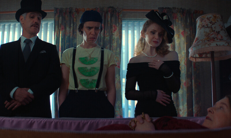Three people stand in a dimly lit room by a window, dressed in funeral attire, looking solemnly at a woman lying in an open casket, creating a somber mood.