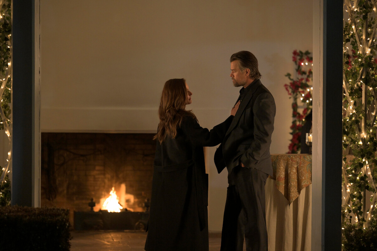 A woman and a man stand facing each other on a decorated patio at night, with string lights, a fireplace burning in the background, and a table covered with a cloth nearby, creating a warm, intimate atmosphere.
