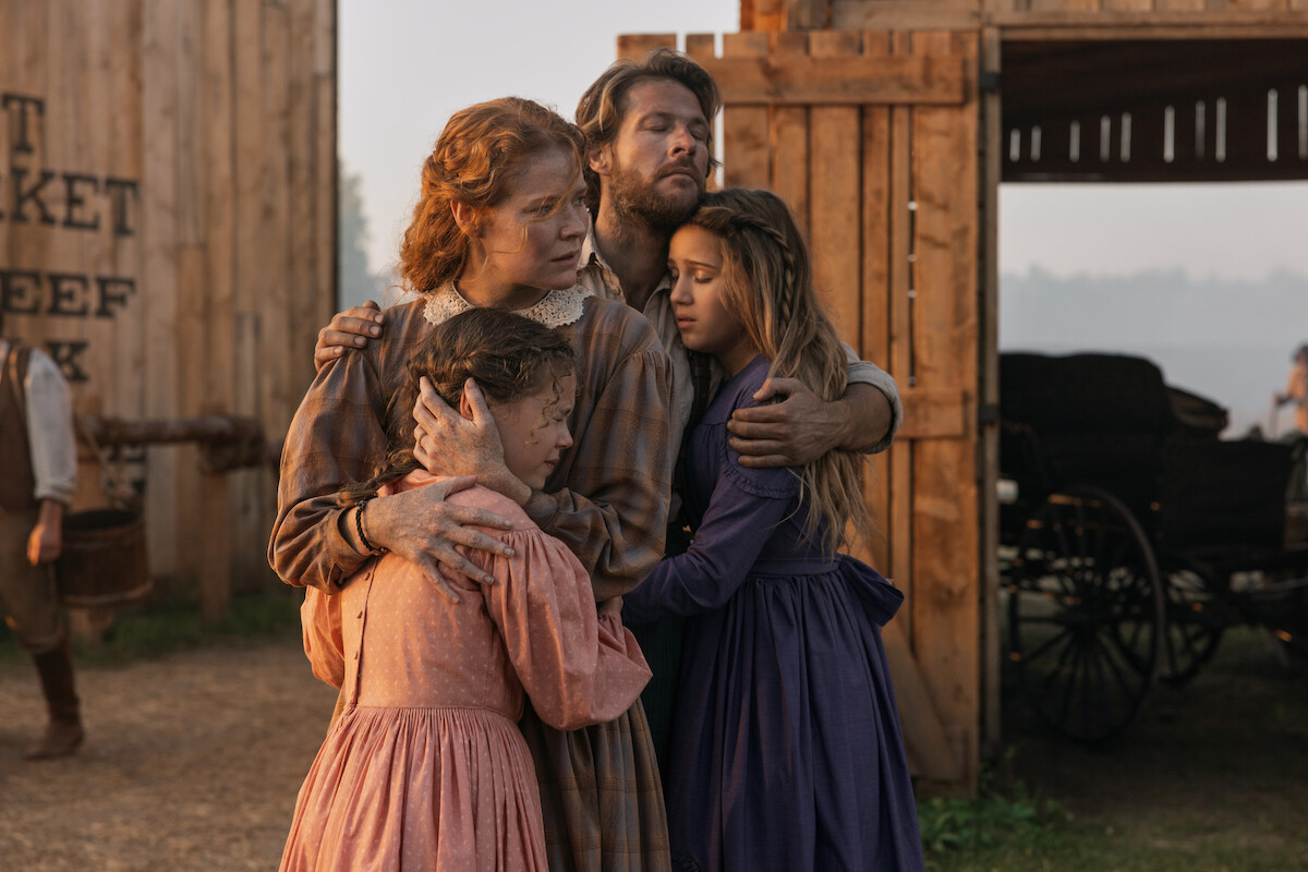 A family of four in historical clothing stands hugging outside wooden barn buildings, looking concerned. The background includes wagon wheels, dirt ground, and faded signage, suggesting a rural or frontier setting in the past.