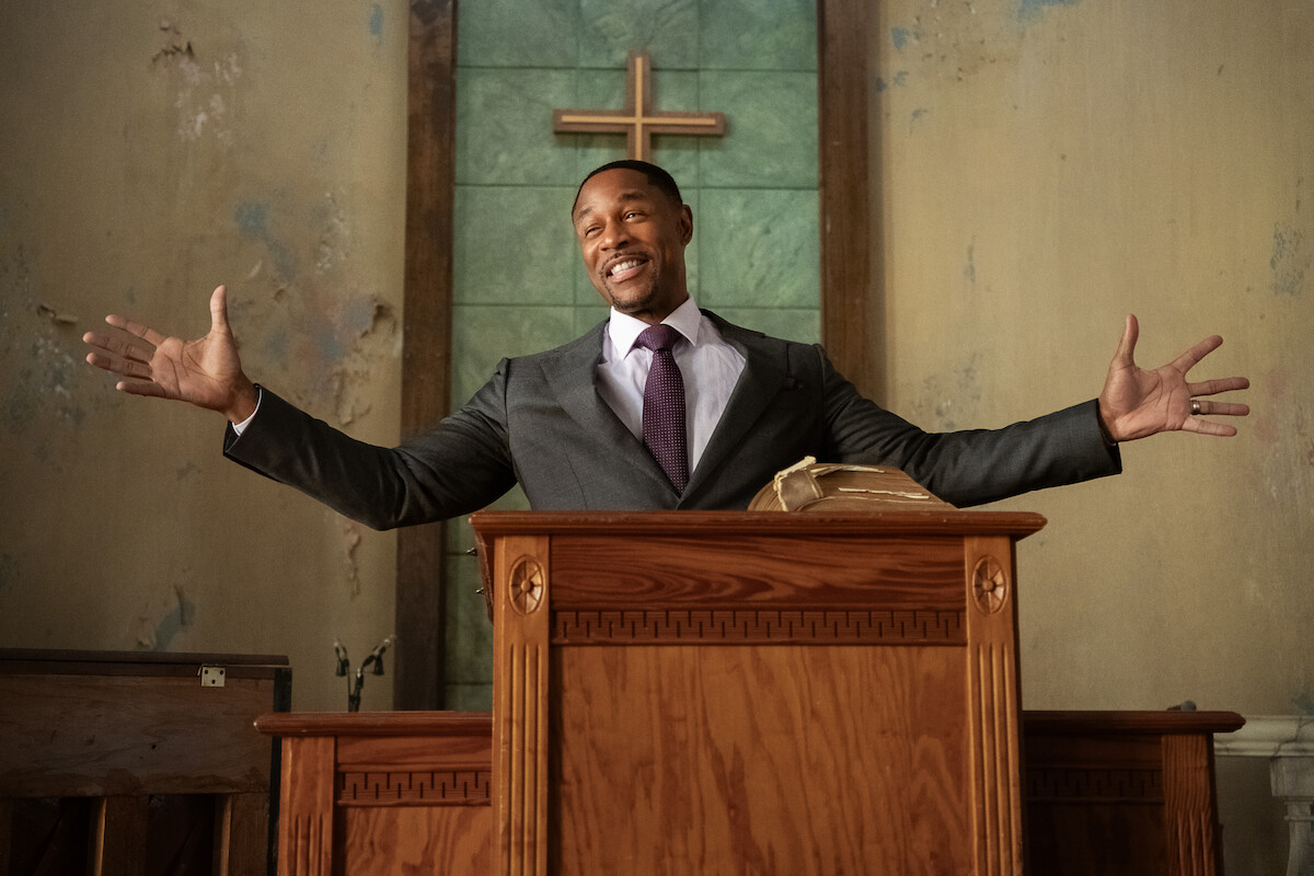Man in a suit standing at a church pulpit, smiling with arms open wide, Bible on podium, wooden cross and worn walls in the background, indoor setting, appears to be giving a sermon or addressing a congregation.