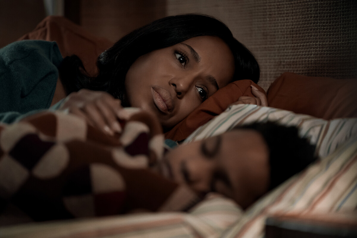 Woman lying in bed looking thoughtful while a child sleeps beside her, both under a patterned blanket in a cozy, softly lit bedroom setting.