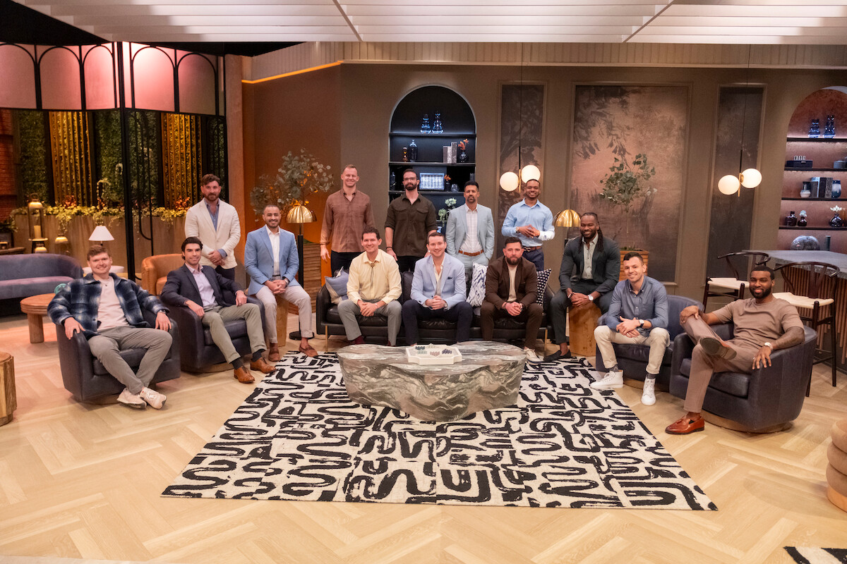 A group of 15 men sit and stand in a stylish, modern lounge with warm lighting, decorative shelves, indoor plants, and abstract art. They pose around a stone coffee table on a patterned rug, creating a relaxed, casual atmosphere.