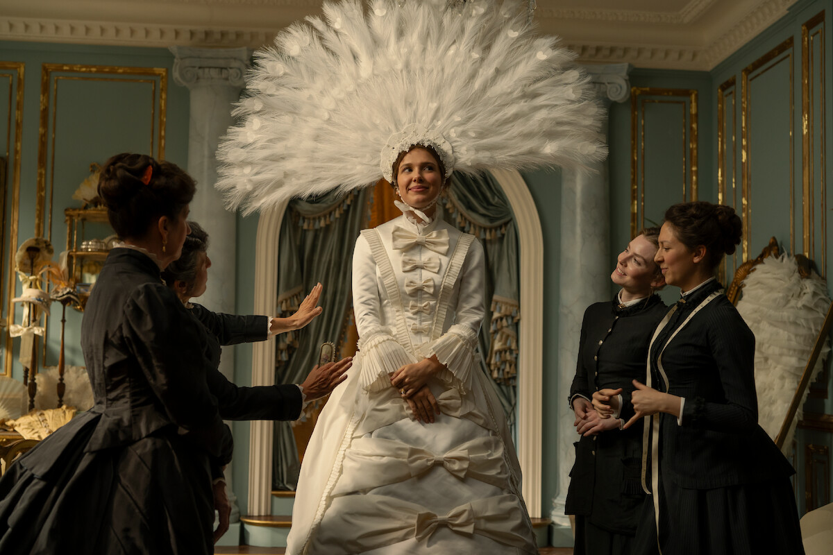 Woman in an elaborate white gown and large feathered headdress stands in an ornate, gold-trimmed room, surrounded by three women dressed in dark period clothing, as they adjust her outfit and admire her.
